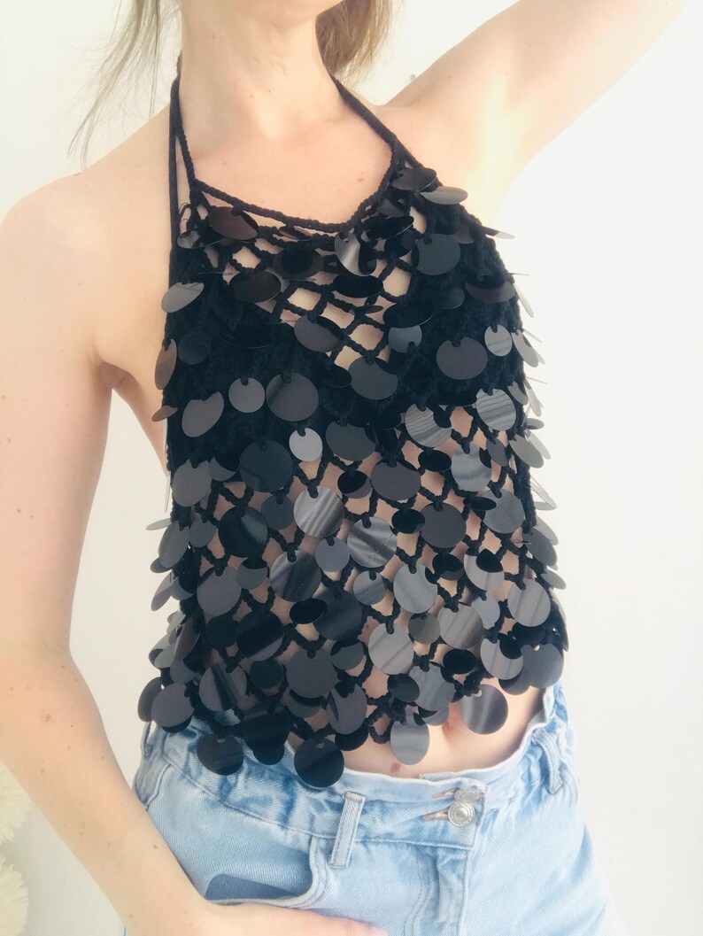 May include: Black halter top with a crochet design and large, round, reflective sequins. The top features a halter neck and a series of sequins cascading down the front. The top is paired with light blue denim jeans.