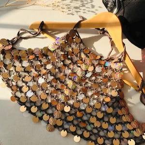 May include: A brown crochet halter top featuring a dense arrangement of circular, iridescent, and metallic gold sequins. The garment is constructed with a fine mesh-like yarn structure and includes thin tie-straps at the neck and back for adjustment.