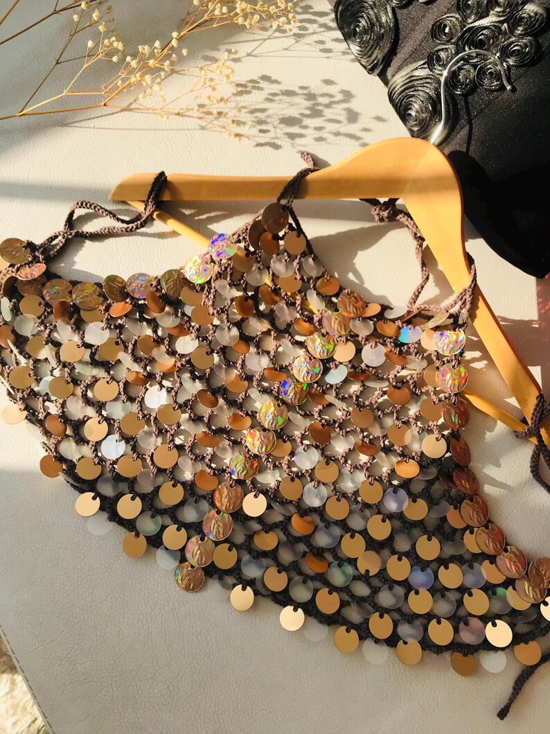 May include: A brown crochet halter top featuring a dense arrangement of circular, iridescent, and metallic gold sequins. The garment is constructed with a fine mesh-like yarn structure and includes thin tie-straps at the neck and back for adjustment.
