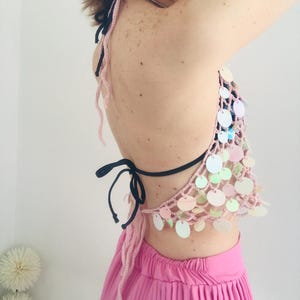 May include: A light pink crochet halter top featuring iridescent circular sequins attached to the mesh-like fabric. The garment includes thin black tie straps at the neck and back, paired with a bright pink pleated skirt worn underneath.