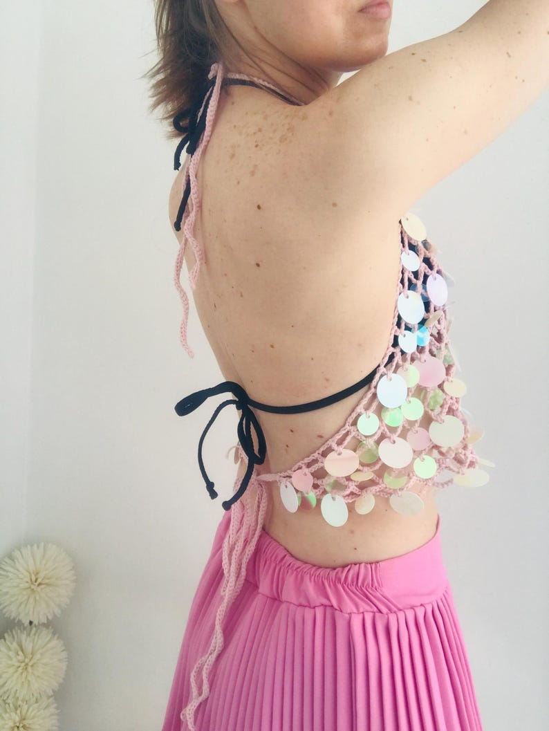 May include: A light pink crochet halter top featuring iridescent circular sequins attached to the mesh-like fabric. The garment includes thin black tie straps at the neck and back, paired with a bright pink pleated skirt worn underneath.