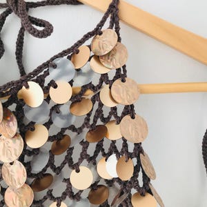 May include: A dark brown crochet halter top featuring a net-like construction adorned with numerous circular metallic discs in shades of gold, bronze, and translucent white. The garment includes braided ties at the neck and a textured open-knit design.