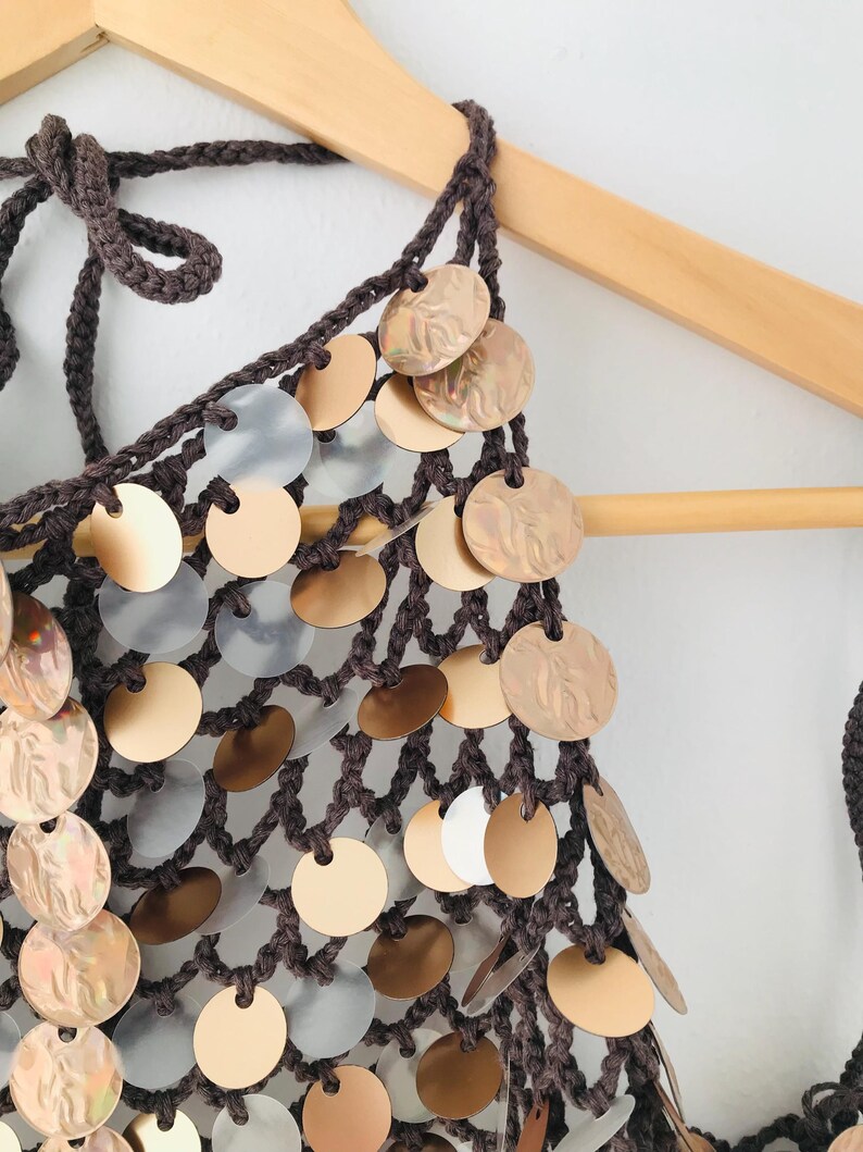 May include: A dark brown crochet halter top featuring a net-like construction adorned with numerous circular metallic discs in shades of gold, bronze, and translucent white. The garment includes braided ties at the neck and a textured open-knit design.
