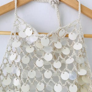 May include: A cream-colored crochet halter top featuring a net-like construction adorned with numerous circular, iridescent white sequins. The garment includes a braided neck tie and is displayed on a wooden hanger against a plain background.
