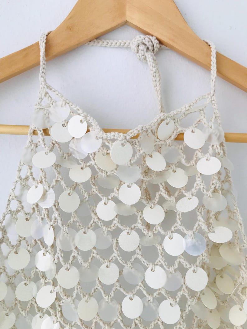 May include: A cream-colored crochet halter top featuring a net-like construction adorned with numerous circular, iridescent white sequins. The garment includes a braided neck tie and is displayed on a wooden hanger against a plain background.