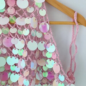 May include: A light pink crochet top featuring a wide-mesh net construction adorned with numerous circular, iridescent sequins. The sequins display shifting colors including white, soft pink, and pale green, attached to the yarn structure.