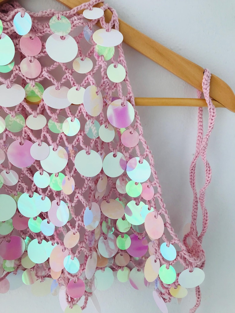 May include: A light pink crochet top featuring a wide-mesh net construction adorned with numerous circular, iridescent sequins. The sequins display shifting colors including white, soft pink, and pale green, attached to the yarn structure.