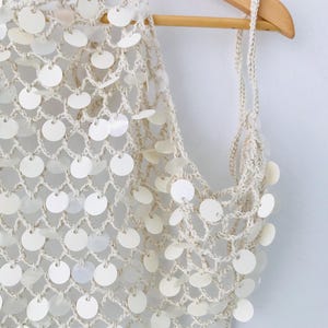 May include: A cream-colored crochet halter top featuring a wide-mesh net pattern adorned with numerous small, circular, iridescent white sequins attached at each intersection of the yarn, suspended from a wooden hanger.