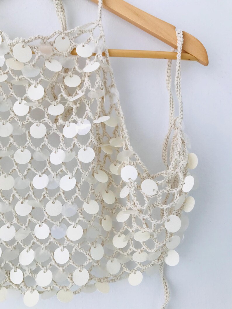 May include: A cream-colored crochet halter top featuring a wide-mesh net pattern adorned with numerous small, circular, iridescent white sequins attached at each intersection of the yarn, suspended from a wooden hanger.
