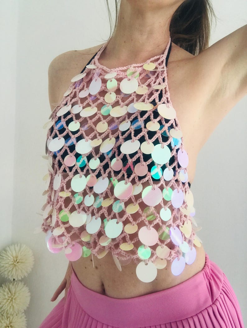 May include: A light pink crochet halter top featuring a wide-gauge net pattern embellished with numerous circular, iridescent sequins that reflect various colors. The garment is sleeveless with a high neckline and an open-back design.