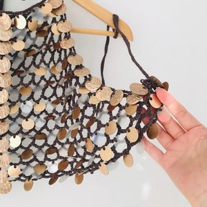 May include: A dark brown crocheted halter top featuring a wide-mesh net construction adorned with numerous circular metallic discs in shades of gold and silver. The garment is suspended from a wooden hanger and features thin crocheted ties.