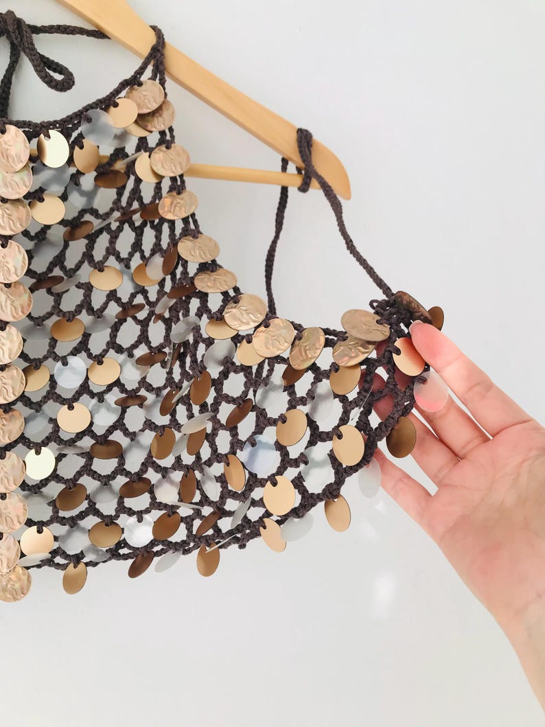 May include: A dark brown crocheted halter top featuring a wide-mesh net construction adorned with numerous circular metallic discs in shades of gold and silver. The garment is suspended from a wooden hanger and features thin crocheted ties.