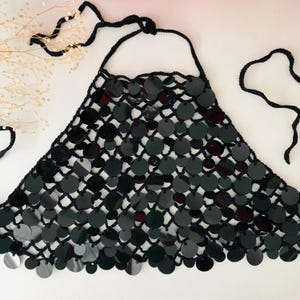 May include: A black halter top with a net design, embellished with many black sequins. The top has a halter neck tie and a triangular shape. The sequins are circular and arranged in a grid pattern, creating a shimmering effect.