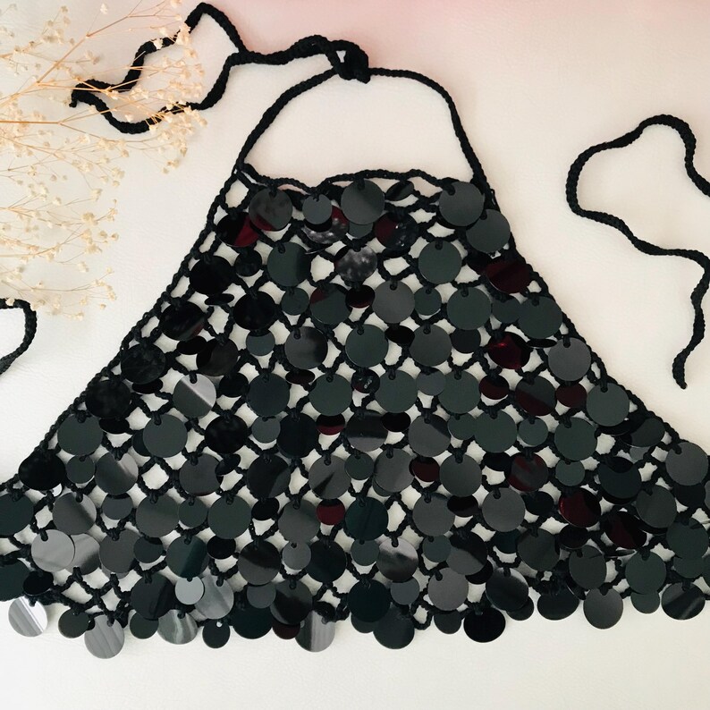 May include: A black halter top with a net design, embellished with many black sequins. The top has a halter neck tie and a triangular shape. The sequins are circular and arranged in a grid pattern, creating a shimmering effect.