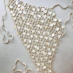 May include: A triangular crochet top featuring an open-knit mesh pattern in a light beige color. The garment is embellished with numerous small, circular, iridescent white sequins attached throughout the netting and includes long tie strings.