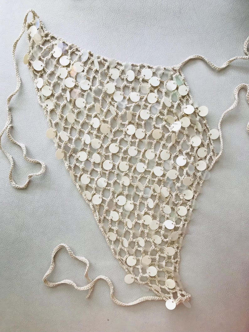 May include: A triangular crochet top featuring an open-knit mesh pattern in a light beige color. The garment is embellished with numerous small, circular, iridescent white sequins attached throughout the netting and includes long tie strings.