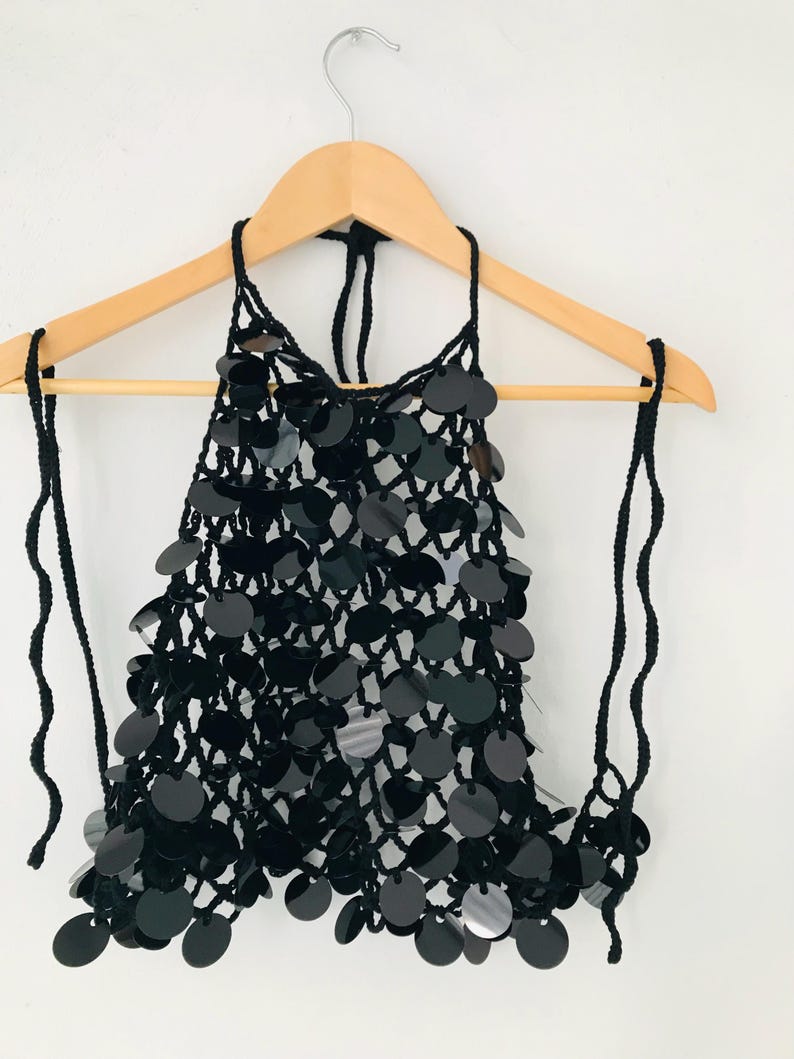 May include: Black halter top with large, round sequins. The top is made of a crocheted net and hangs on a wooden hanger. The halter straps are made of black yarn. The sequins are shiny and reflect light.