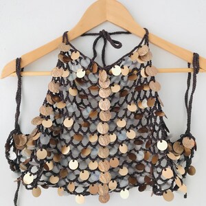 May include: A dark brown crochet halter top featuring a net-like structure adorned with numerous circular metallic sequins in shades of gold, bronze, and silver. The garment includes thin crocheted ties at the neck and sides for adjustable closure.