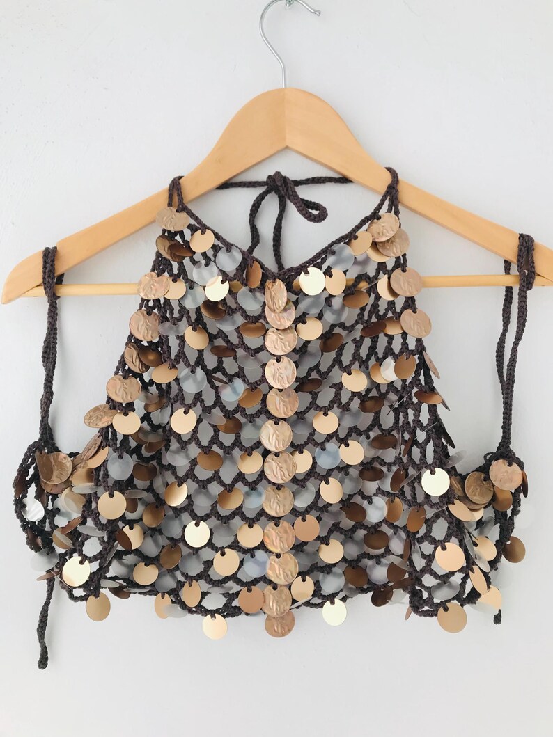 May include: A dark brown crochet halter top featuring a net-like structure adorned with numerous circular metallic sequins in shades of gold, bronze, and silver. The garment includes thin crocheted ties at the neck and sides for adjustable closure.