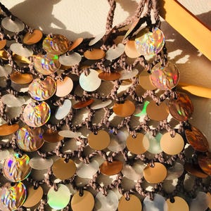 May include: A brown crocheted net top featuring a dense arrangement of circular sequins. The sequins alternate between solid metallic bronze, translucent white, and iridescent discs that reflect light with a multi-colored, holographic sheen.
