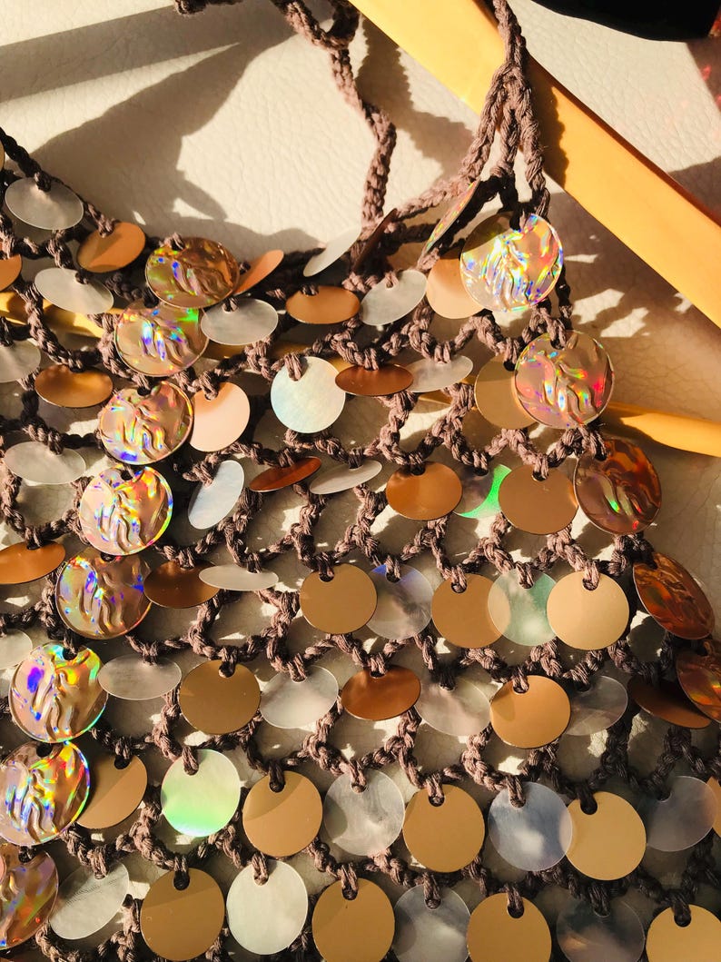 May include: A brown crocheted net top featuring a dense arrangement of circular sequins. The sequins alternate between solid metallic bronze, translucent white, and iridescent discs that reflect light with a multi-colored, holographic sheen.