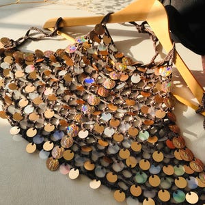 May include: A dark brown crochet halter top featuring a mesh construction adorned with numerous circular, iridescent, and metallic gold sequins. The garment is suspended on a wooden hanger and includes thin braided ties at the neck and back.