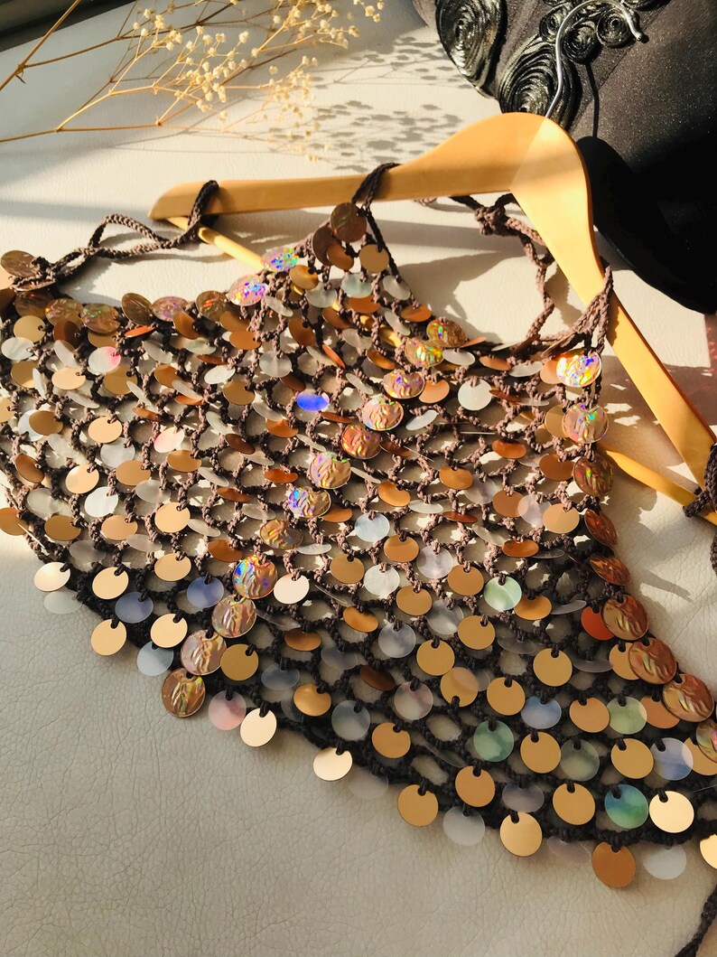 May include: A dark brown crochet halter top featuring a mesh construction adorned with numerous circular, iridescent, and metallic gold sequins. The garment is suspended on a wooden hanger and includes thin braided ties at the neck and back.