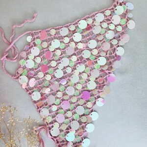 May include: A triangular crochet top crafted from light pink yarn, featuring an open-knit mesh pattern adorned with numerous iridescent, circular sequins that reflect various colors. The garment includes thin, matching pink tie-strings at the corners.