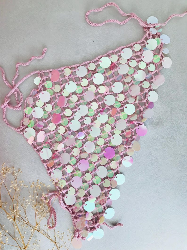 May include: A triangular crochet top crafted from light pink yarn, featuring an open-knit mesh pattern adorned with numerous iridescent, circular sequins that reflect various colors. The garment includes thin, matching pink tie-strings at the corners.