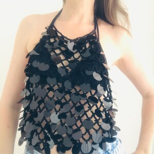 May include: A black halter top with a net-like design, decorated with many black sequins. The top is handmade and appears to be crocheted, offering a unique texture and style. The top is paired with blue jeans.