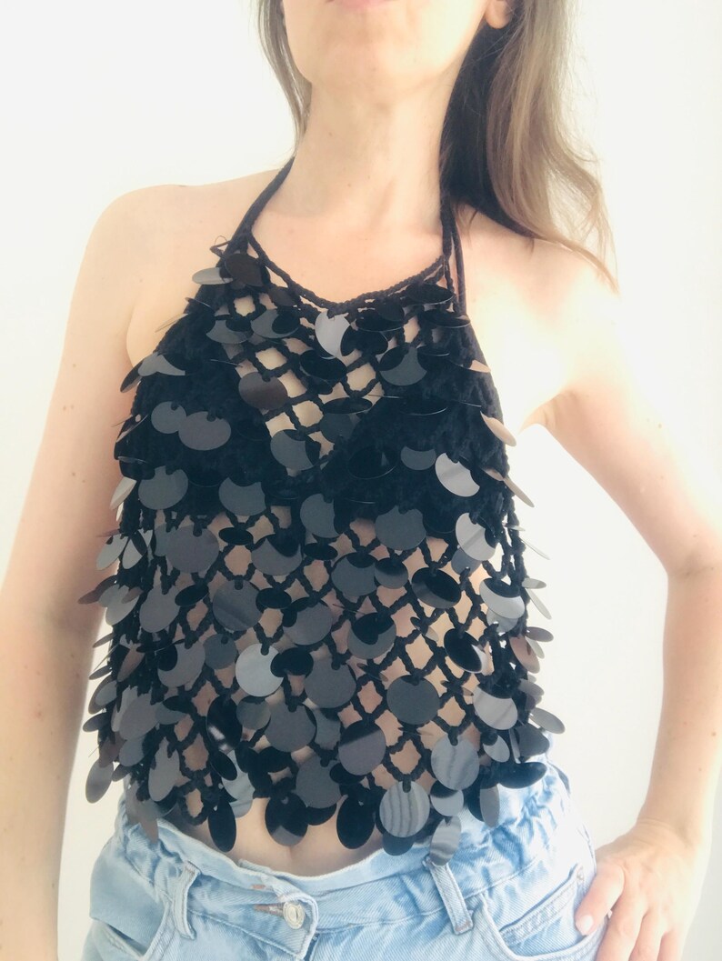 May include: A black halter top with a net-like design, decorated with many black sequins. The top is handmade and appears to be crocheted, offering a unique texture and style. The top is paired with blue jeans.