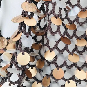 May include: A dark brown crocheted net bag featuring a dense pattern of circular metallic discs. The discs alternate between a matte gold finish and a translucent, iridescent white material, each securely attached to the knotted yarn structure.