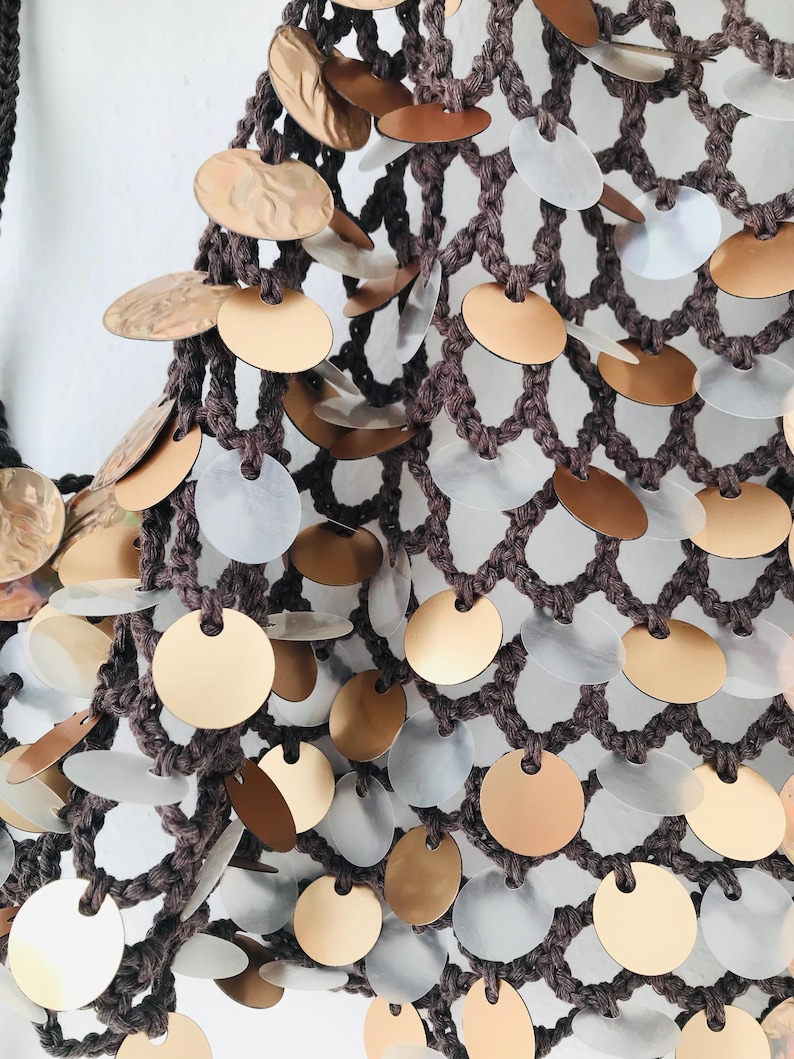 May include: A dark brown crocheted net bag featuring a dense pattern of circular metallic discs. The discs alternate between a matte gold finish and a translucent, iridescent white material, each securely attached to the knotted yarn structure.