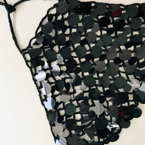 May include: A black sequin top with a halter neck. The top features a crocheted net base, with large, round, black sequins attached. The sequins are reflective. The top is a triangle shape and is designed to be worn around the neck.