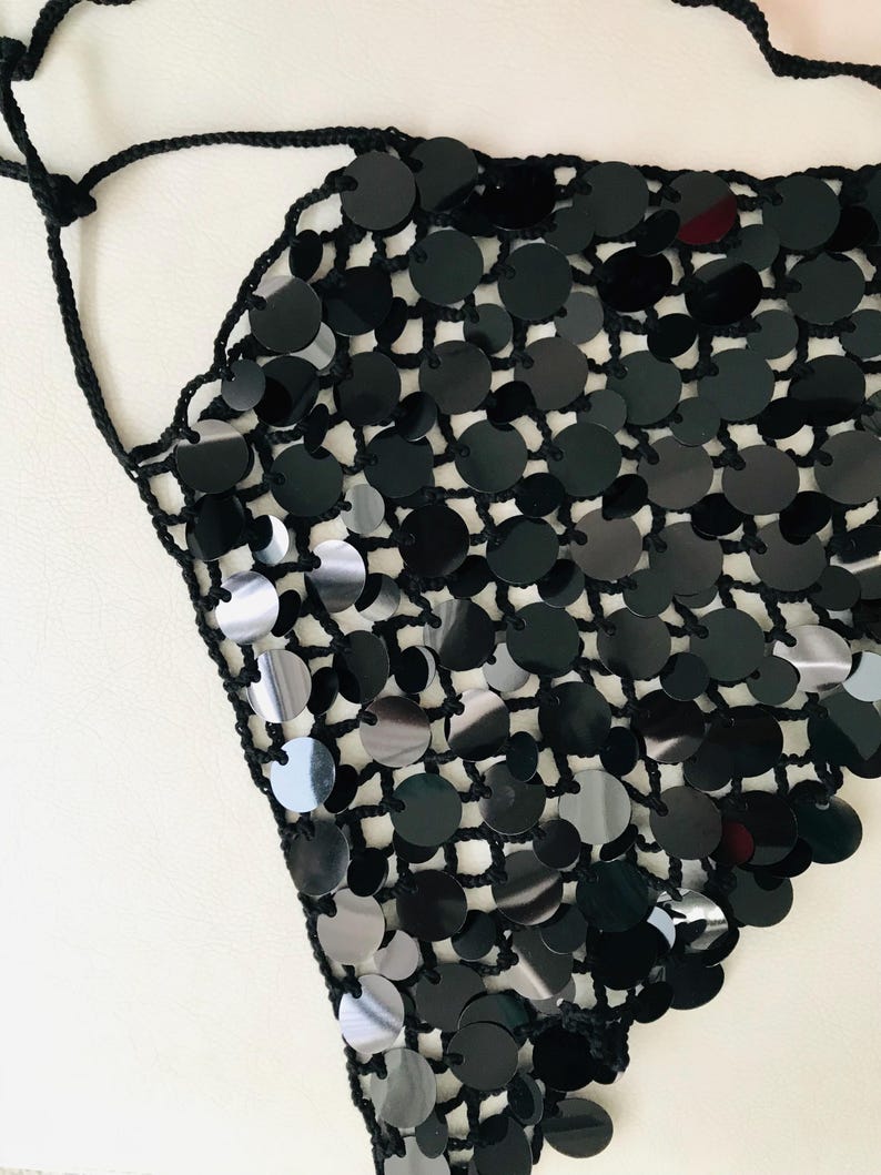 May include: A black sequin top with a halter neck. The top features a crocheted net base, with large, round, black sequins attached. The sequins are reflective. The top is a triangle shape and is designed to be worn around the neck.