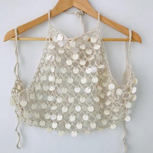 May include: A cream-colored crochet halter top featuring a net-like pattern adorned with numerous circular, iridescent white sequins. The garment is suspended from a wooden hanger and includes thin, adjustable crochet ties at the neck and sides.