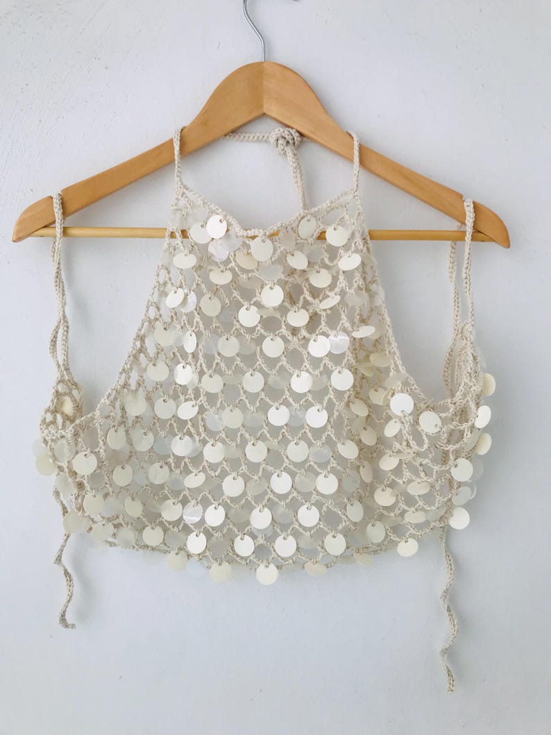 May include: A cream-colored crochet halter top featuring a net-like pattern adorned with numerous circular, iridescent white sequins. The garment is suspended from a wooden hanger and includes thin, adjustable crochet ties at the neck and sides.