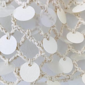 May include: A cream-colored macrame net featuring a grid of knotted cord. Attached to the intersections of the cord are numerous circular, semi-translucent white discs that hang freely, creating a layered and textured appearance across the surface.