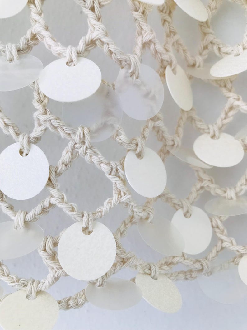May include: A cream-colored macrame net featuring a grid of knotted cord. Attached to the intersections of the cord are numerous circular, semi-translucent white discs that hang freely, creating a layered and textured appearance across the surface.