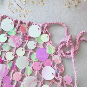 May include: A light pink crochet top featuring a net-like structure adorned with numerous circular, iridescent sequins. The garment includes thin, matching pink crocheted ties at the sides for fastening, creating a textured and reflective appearance.