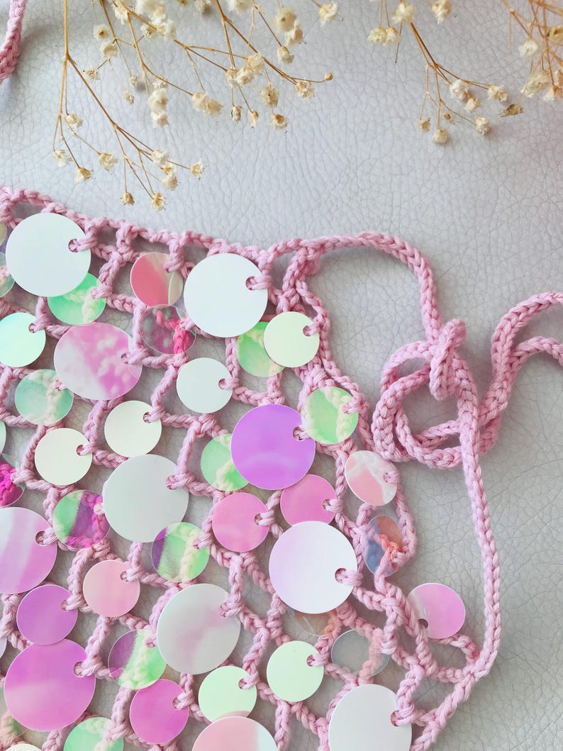 May include: A light pink crochet top featuring a net-like structure adorned with numerous circular, iridescent sequins. The garment includes thin, matching pink crocheted ties at the sides for fastening, creating a textured and reflective appearance.