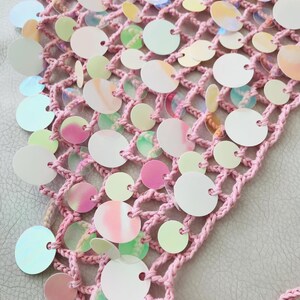 May include: A light pink crocheted net fabric adorned with numerous circular, iridescent sequins. The sequins feature a reflective, pearlescent finish that shifts in color, attached to the open-weave yarn structure in varying sizes across the surface.