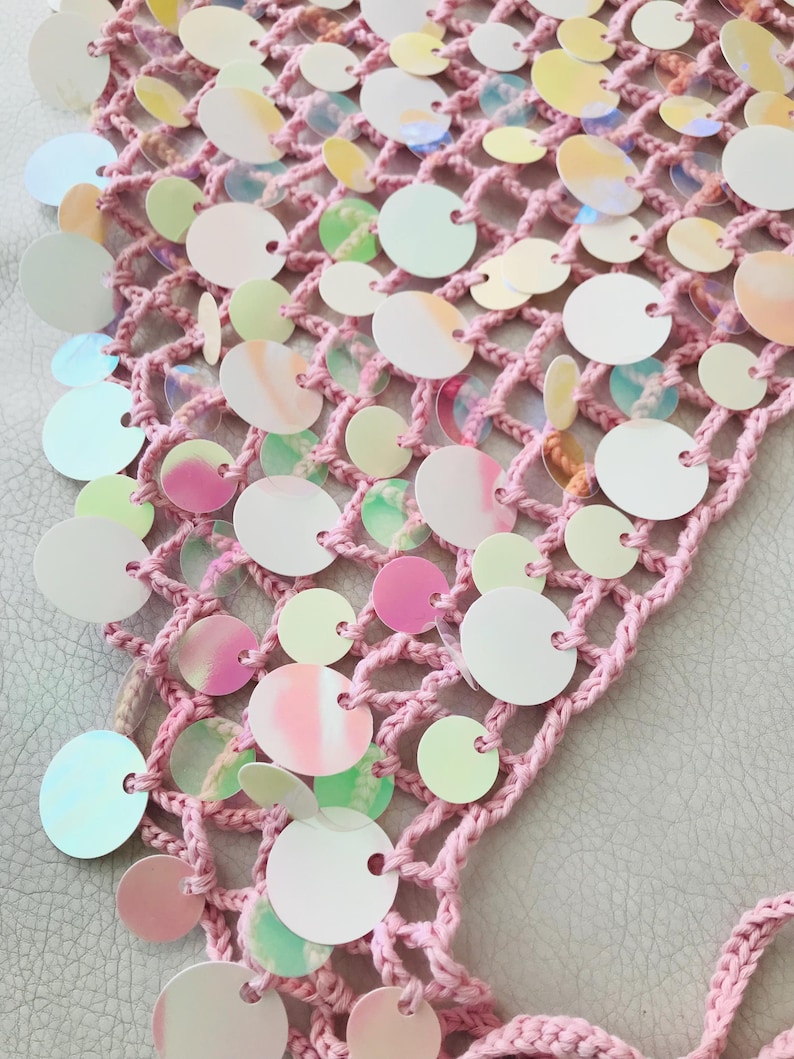 May include: A light pink crocheted net fabric adorned with numerous circular, iridescent sequins. The sequins feature a reflective, pearlescent finish that shifts in color, attached to the open-weave yarn structure in varying sizes across the surface.