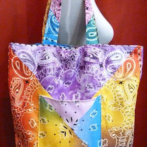 May include: A tote bag constructed from patchwork fabric featuring a vibrant paisley bandana print in shades of purple, orange, yellow, blue, and green. The bag includes two shoulder straps and a single exterior pocket on the front panel.
