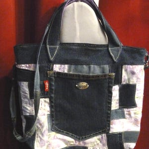 May include: A patchwork tote bag constructed from dark blue denim and light purple floral fabric. The bag features a central denim pocket with a Wrangler brand tag and a metallic oval logo plate. It includes two top handles and a shoulder strap.