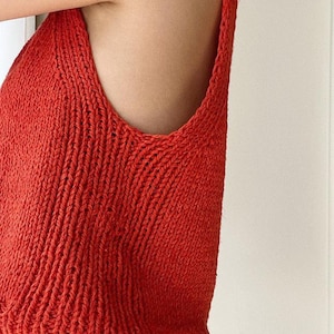 Puede incluir: Primer plano de una camiseta sin mangas de punto rojo vibrante. La parte superior presenta un cuello redondo y tirantes anchos, mostrando un patrón de punto texturizado. La prenda está hecha de un material suave y transpirable, perfecto para el clima cálido.