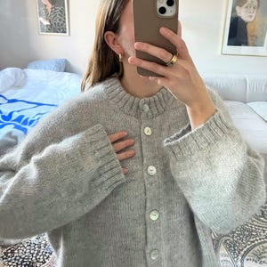 Op de afbeelding: Een lichtgrijze cardigan met knopen, een ronde hals en lange mouwen. De trui heeft grote, ronde, witte knopen en een geribbelde kraag en manchetten. Het materiaal lijkt zachte wol of een wolmix te zijn.