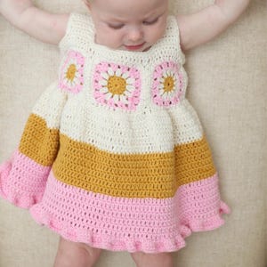 Op de afbeelding: Een handgemaakte gehaakte babyjurk in crème, mosterdgeel en roze. De jurk heeft een vierkante halslijn en klokmouwen. De lijfje heeft drie vierkante bloemmotieven. De rok heeft een gefranjerde zoom.