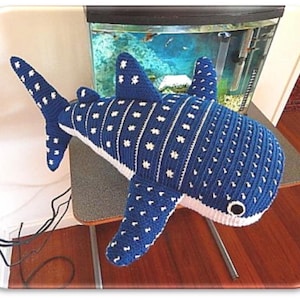 Op de afbeelding: Een grote, handgemaakte gehaakte walvishaai kussen in blauw en wit. Het kussen heeft een gedetailleerd patroon van witte stippen en strepen op een blauwe achtergrond. De vinnen en staart zijn ook blauw met witte accenten. Het kussen ligt op een houten oppervlak.