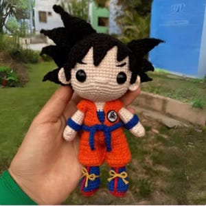 Op de afbeelding: Gehaakte pop van een personage met stekelig zwart haar, gekleed in een oranje en blauwe outfit. De pop heeft een rond gezicht, zwarte ogen en een symbool op de borst. De schoenen van de pop zijn blauw met gele strikken.